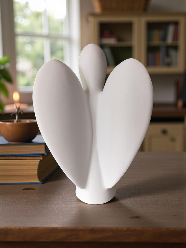 May include: A white ceramic angel sculpture with outstretched wings. The sculpture has a smooth, matte finish and is displayed on a wooden surface. The angel's wings are large and curved, creating a heart-like shape.