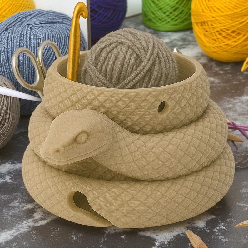 May include: A tan-colored snake-shaped yarn bowl with a ball of yarn inside. A gold crochet hook and scissors are visible. The bowl has a textured, scale-like pattern. Other yarn balls in blue, purple, green, and yellow are in the background.