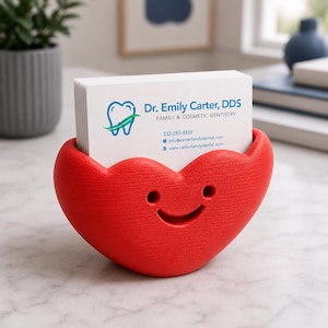 May include: A red heart-shaped business card holder with a smiling face design. The holder contains white business cards with the text "Dr. Emily Carter, DDS" and contact information. The holder is on a white surface.