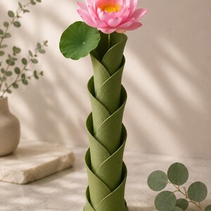 May include: A green, spiral-shaped vase holds a pink lotus flower with a yellow center and a green leaf. The vase has a textured surface and a unique, layered design, creating a modern and elegant look. The flower is in full bloom.