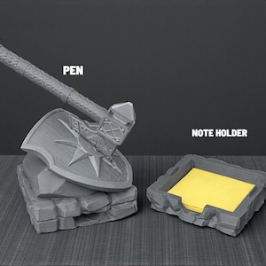 May include: A gray pen holder shaped like a hammer and shield, with the word "PEN" above it. Beside it is a matching gray note holder with yellow sticky notes and the words "NOTE HOLDER".