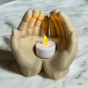 Puede incluir: Un portavelas beige con forma de dos manos que sostienen una vela de t&eacute; blanca con una llama amarilla parpadeante. El portavelas est&aacute; hecho de un material liso y descansa sobre una superficie de m&aacute;rmol.