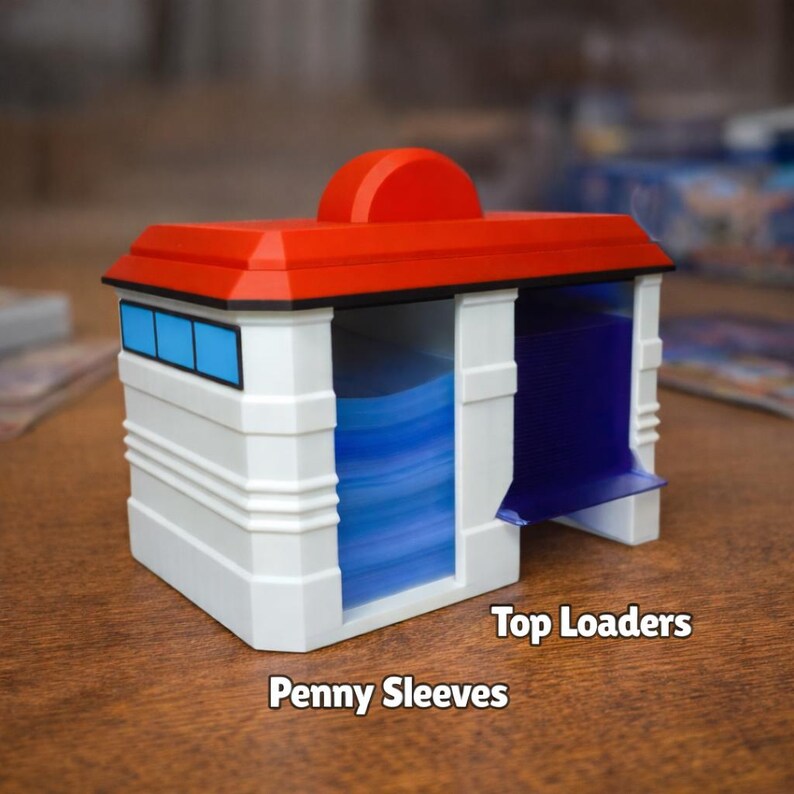 Puede incluir: Un contenedor de almacenamiento de tarjetas blanco y rojo con detalles azules. El contenedor tiene un techo rojo y un &aacute;rea de visualizaci&oacute;n azul. Los textos "Top Loaders" y "Penny Sleeves" son visibles.