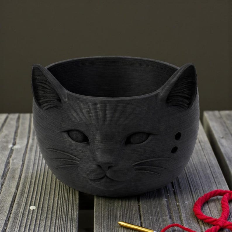 May include: A black cat-shaped planter with a textured finish. The planter has a detailed cat face with eyes, nose, and whiskers. The ears are upright, and there are two small holes on one side. It rests on a wooden surface.