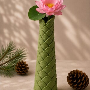 May include: A green, textured vase with a lotus flower and leaf. The pink flower has a yellow center. The vase has a scale-like pattern. Pine cones and a pine branch are in the background.