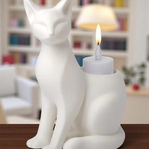Op de afbeelding: Een witte keramische kattenfiguur met een kandelaar. De kat zit met gesloten ogen. Een kleine witte kaars met een brandende vlam staat in een houder op de rug van de kat. De figuur staat op een bruin oppervlak.