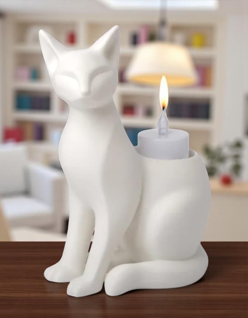 Op de afbeelding: Een witte keramische kattenfiguur met een kandelaar. De kat zit met gesloten ogen. Een kleine witte kaars met een brandende vlam staat in een houder op de rug van de kat. De figuur staat op een bruin oppervlak.