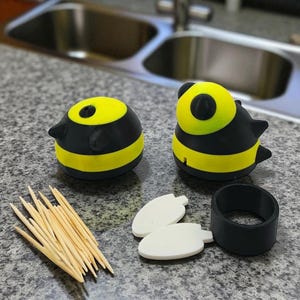 May include: Two bee-shaped toothpick holders with yellow and black stripes, along with a pile of toothpicks, two white leaf-shaped pieces, and a black ring. The holders have a round shape with a yellow top and black accents.