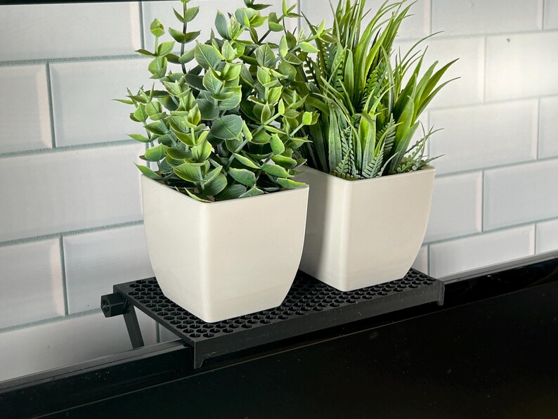 May include: Two potted plants, one with green leaves and one with long green blades, sit on a black metal shelf attached to a black surface. The shelf has a grid pattern.