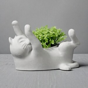 May include: A white ceramic cat-shaped planter with its paws raised, holding a small green plant. The cat is lying on its back, with a smiling expression. The planter is a decorative item for home decor.