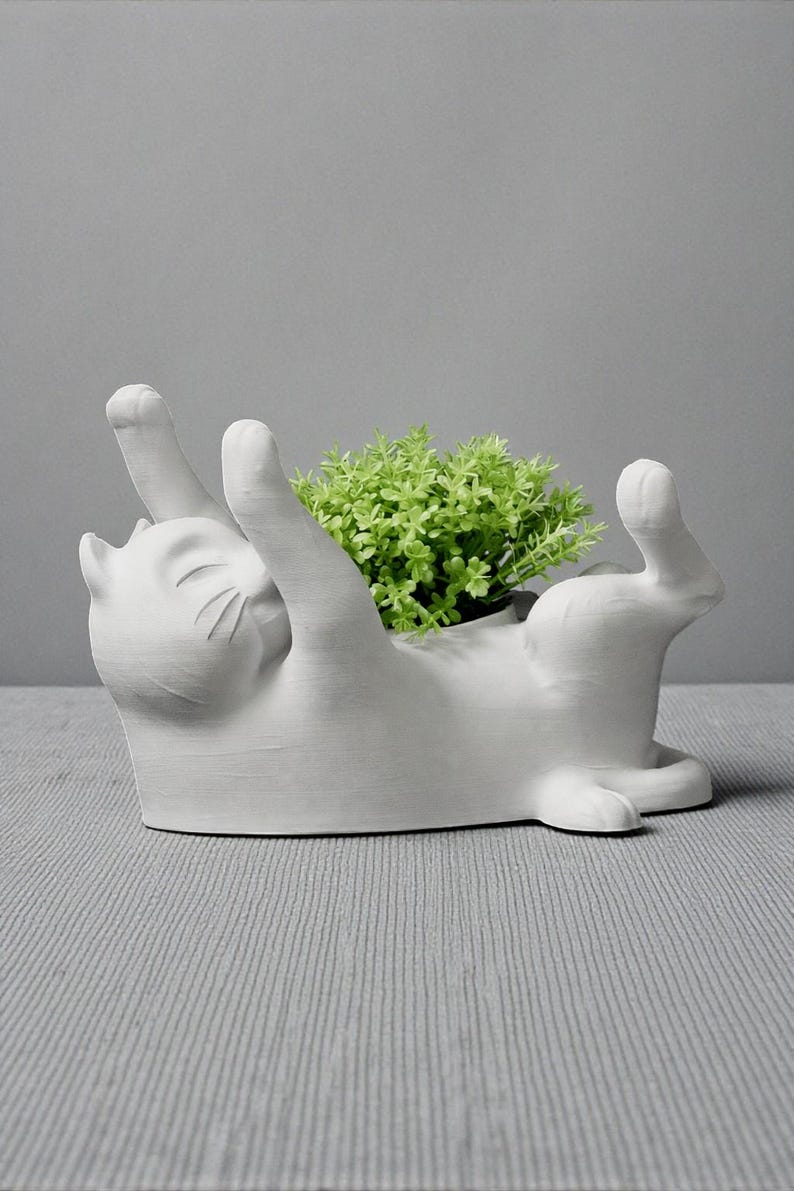 May include: A white ceramic cat-shaped planter with its paws raised, holding a small green plant. The cat is lying on its back, with a smiling expression. The planter is a decorative item for home decor.