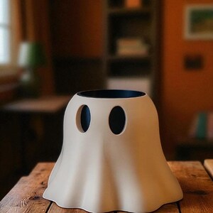 May include: A beige ghost-shaped ceramic vase with two oval eye cutouts and a black interior. The vase sits on a wooden table, with a blurred background of a room.