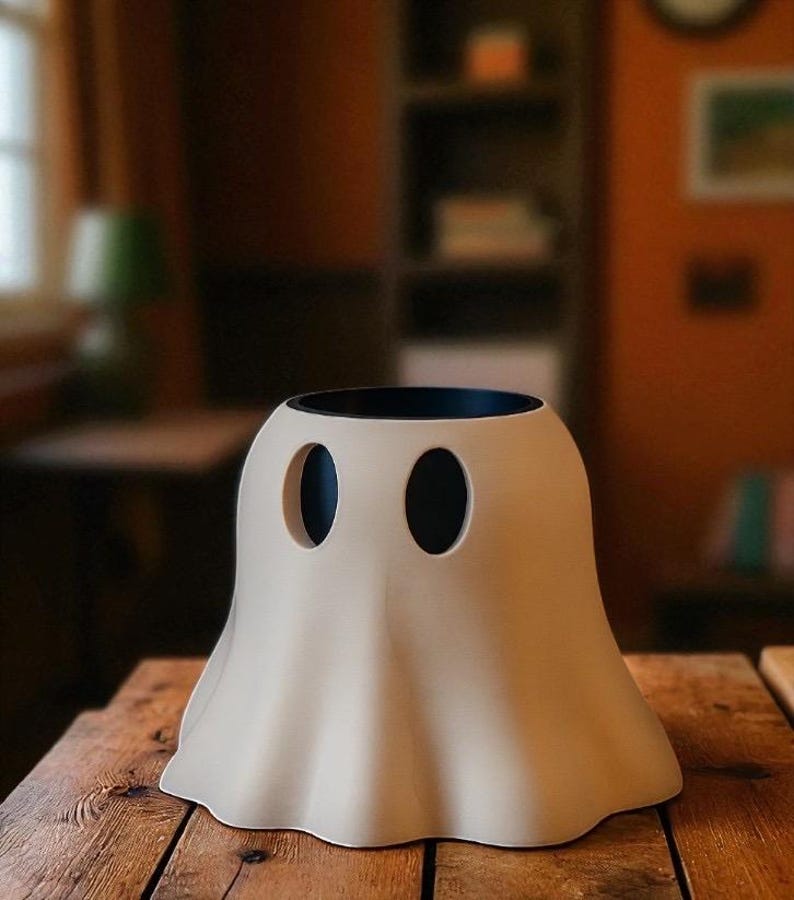 May include: A beige ghost-shaped ceramic vase with two oval eye cutouts and a black interior. The vase sits on a wooden table, with a blurred background of a room.