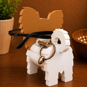 May include: A dog-shaped eyeglasses and key holder in white and brown. The holder features a brown head and ears, a white body, and holds black eyeglasses and a key ring. The wooden holder sits on a wooden surface.