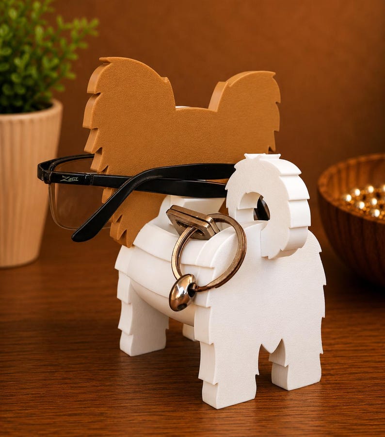 May include: A dog-shaped eyeglasses and key holder in white and brown. The holder features a brown head and ears, a white body, and holds black eyeglasses and a key ring. The wooden holder sits on a wooden surface.