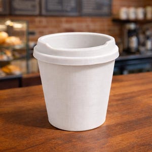 May include: A white disposable coffee cup with a lid. The cup has a textured surface and is sitting on a wooden table. The background includes a brick wall and a display case.