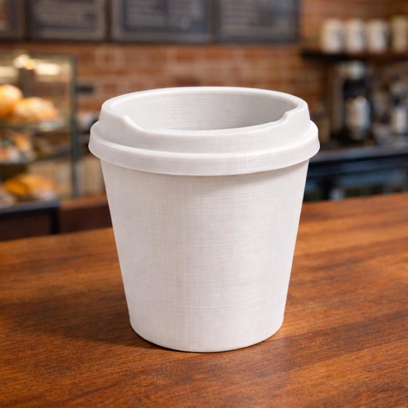 May include: A white disposable coffee cup with a lid. The cup has a textured surface and is sitting on a wooden table. The background includes a brick wall and a display case.