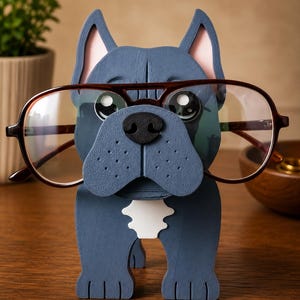 May include: A blue wooden bulldog-shaped glasses holder with brown-framed glasses. The holder has pink inner ears, a black nose, and a white patch on its chest. The glasses are positioned as if the bulldog is wearing them.