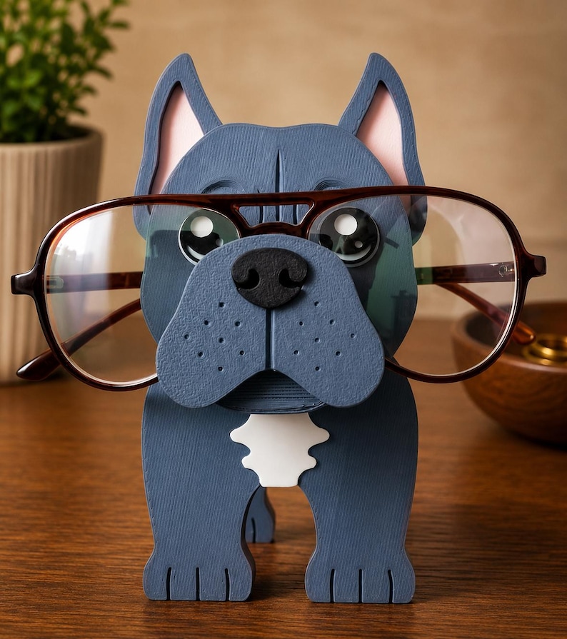 May include: A blue wooden bulldog-shaped glasses holder with brown-framed glasses. The holder has pink inner ears, a black nose, and a white patch on its chest. The glasses are positioned as if the bulldog is wearing them.