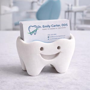 Op de afbeelding: Een witte, tandvormige visitekaarthouder met een lachend gezicht. De houder bevat visitekaartjes voor Dr. Emily Carter, DDS, Family & Cosmetic Dentistry. De kaarten hebben een blauw en groen tandlogo.