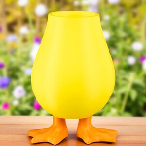 Puede incluir: Un jarr&oacute;n amarillo brillante con forma de huevo y patas de pato naranjas. El jarr&oacute;n tiene una abertura ancha y un cuerpo liso y redondeado. Las patas de pato son detalladas y a&ntilde;aden un toque juguet&oacute;n. El fondo es un jard&iacute;n borroso.