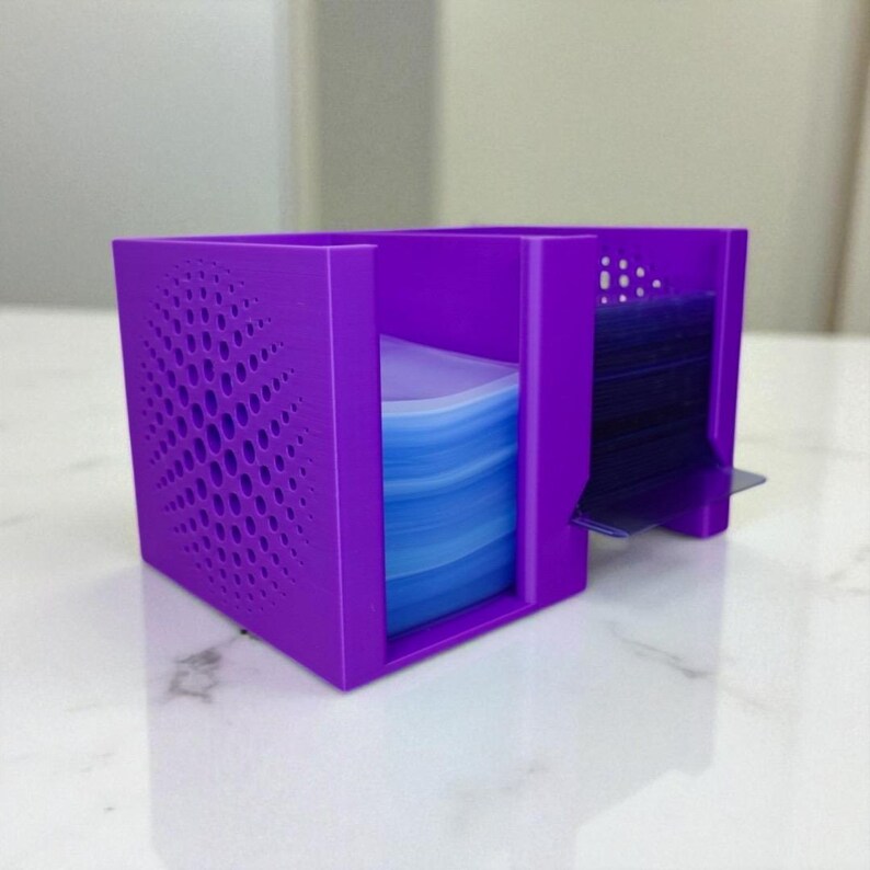 May include: A purple card holder with a geometric design on one side. The holder has two compartments, one filled with blue cards and the other with dark cards. The holder is made of plastic and sits on a white surface.