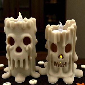 May include: Two off-white Halloween-themed candle decorations. Each candle has a skull face design with eye and mouth cutouts. The candles have a melted wax appearance and are lit with a flickering LED light.