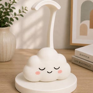 K&ouml;nnte beinhalten: Eine wei&szlig;e, wolkenf&ouml;rmige Schreibtischlampe mit einem l&auml;chelnden Gesicht und rosa Wangen. Die Lampe hat einen gebogenen Arm und steht auf einem runden Sockel. Die Lampe steht auf einer Holzoberfl&auml;che.