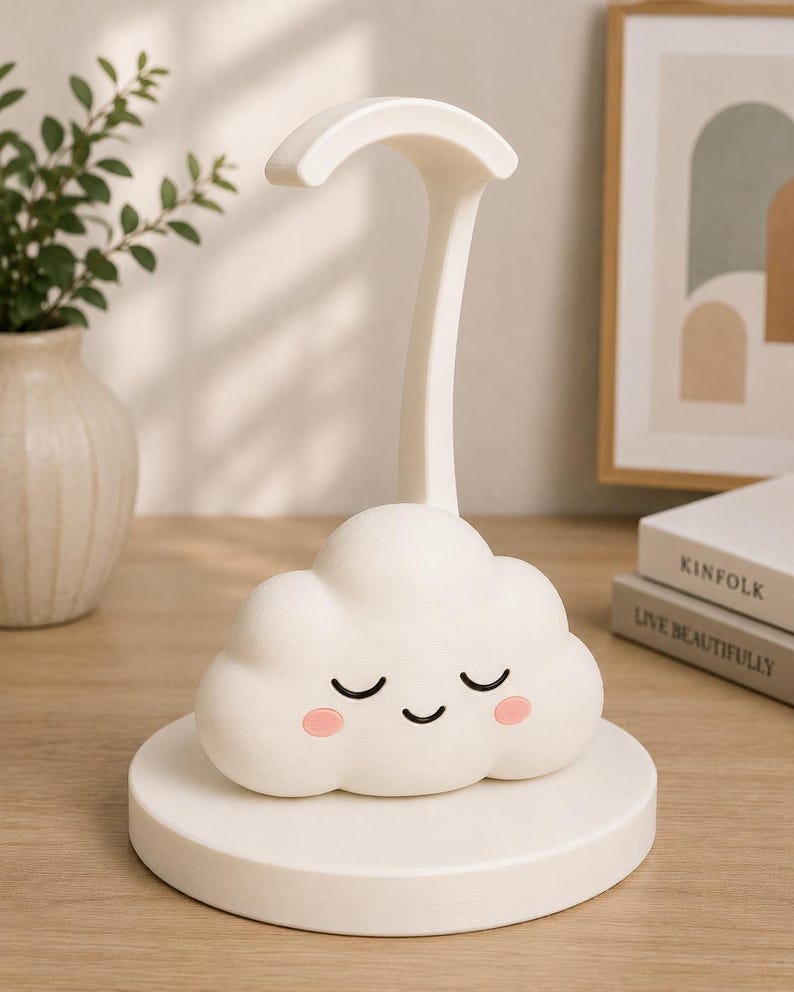 K&ouml;nnte beinhalten: Eine wei&szlig;e, wolkenf&ouml;rmige Schreibtischlampe mit einem l&auml;chelnden Gesicht und rosa Wangen. Die Lampe hat einen gebogenen Arm und steht auf einem runden Sockel. Die Lampe steht auf einer Holzoberfl&auml;che.
