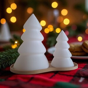 May include: Two white, frosted Christmas tree-shaped decorations on a wooden tray. The trees are different sizes, with a larger one on the left and a smaller one on the right. The background has blurred lights and a red plaid tablecloth.