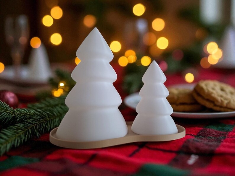 May include: Two white, frosted Christmas tree-shaped decorations on a wooden tray. The trees are different sizes, with a larger one on the left and a smaller one on the right. The background has blurred lights and a red plaid tablecloth.