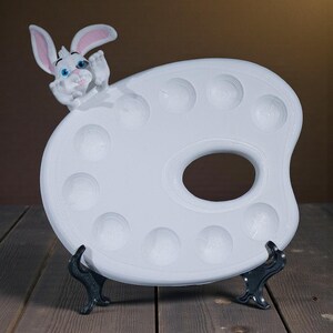 May include: A white ceramic artist's palette with a cartoon bunny figure. The palette has multiple circular wells and a thumb hole. The bunny has pink-lined ears and blue eyes. The palette is supported by a black stand, set against a brown background.