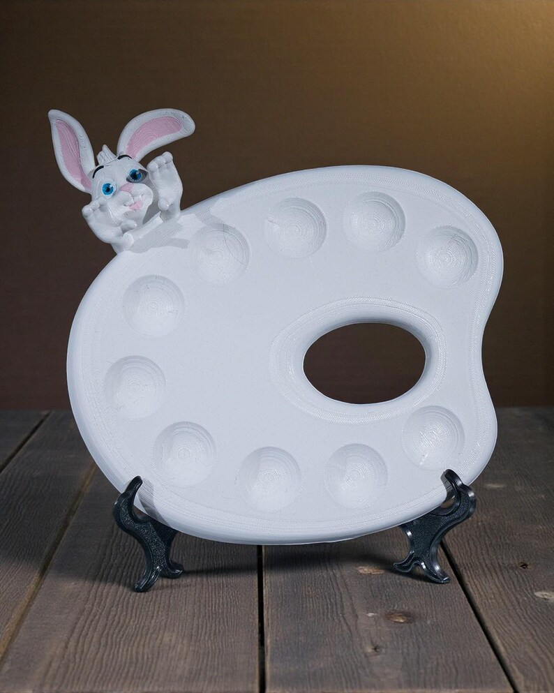 May include: A white ceramic artist's palette with a cartoon bunny figure. The palette has multiple circular wells and a thumb hole. The bunny has pink-lined ears and blue eyes. The palette is supported by a black stand, set against a brown background.