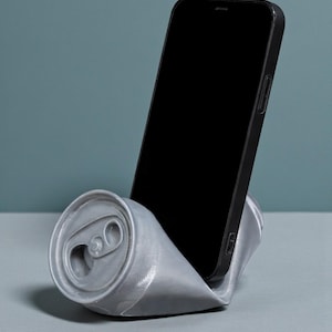 May include: A silver-colored phone stand shaped like a crushed soda can. A black smartphone is placed in the stand, with the screen facing forward. The stand is on a gray surface, with a teal background.