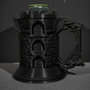 May include: Black castle-shaped mug with a handle featuring a dragon design. The mug has a cylindrical body with arched openings, each displaying the number "5". A green and silver can is inside the mug. The mug is made of a dark material.