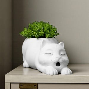 May include: A white cat-shaped planter with a green plant inside. The cat is lying down with its mouth open, as if yawning. The planter is made of ceramic or a similar material and sits on a light-colored surface.