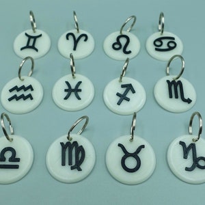 May include: Twelve white round keyrings with black zodiac symbols. The symbols are: Gemini, Aries, Leo, Cancer, Aquarius, Pisces, Sagittarius, Scorpio, Libra, Virgo, Taurus, and Capricorn.