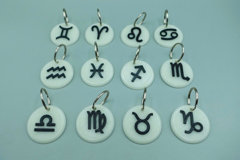 May include: Twelve white round keyrings with black zodiac symbols. The symbols are: Gemini, Aries, Leo, Cancer, Aquarius, Pisces, Sagittarius, Scorpio, Libra, Virgo, Taurus, and Capricorn.