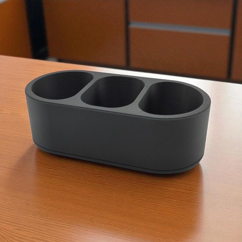 May include: A matte black desk organizer with three oval compartments. The organizer is made of a solid material and sits on a wooden surface. The design is simple and functional, suitable for office or home use.