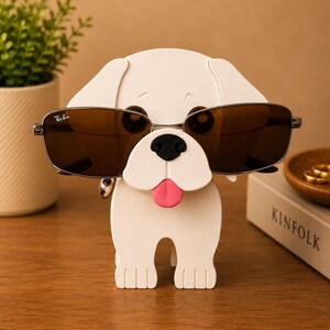 May include: A white dog-shaped eyeglass holder with a black nose and a pink tongue. The holder is wearing a pair of brown-tinted sunglasses. The sunglasses are held in place by the dog's head. The holder is on a wooden surface.