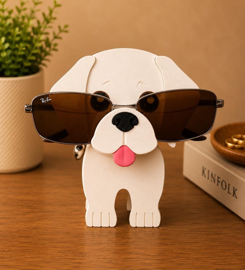 May include: A white dog-shaped eyeglass holder with a black nose and a pink tongue. The holder is wearing a pair of brown-tinted sunglasses. The sunglasses are held in place by the dog's head. The holder is on a wooden surface.