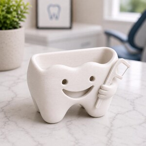 May include: A white ceramic tooth-shaped container with a smiling face and holding a dental floss pick. The container has a rectangular opening at the top and is designed to hold small items. The background includes a framed picture of a tooth.