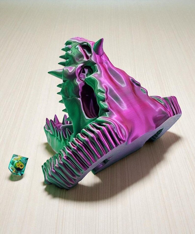 Op de afbeelding: Een 3D-geprinte, veelkleurige drakenkop met een gedetailleerd ontwerp. De schedel is voornamelijk groen en paars, met scherpe tanden en hoorns. Een kleine, veelkleurige dobbelsteen ligt naast de schedel. Het is een decoratief item.