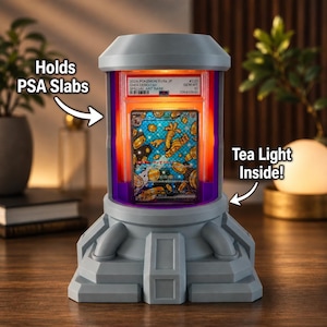 Op de afbeelding: Een grijze display met een theelichtje binnenin, ontworpen om PSA-slabs te bevatten. De behuizing heeft een cilindrisch ontwerp met een paars en oranje verlicht interieur, met een Pok&eacute;mon-kaart. Tekst op de behuizing luidt "Holds PSA Slabs" en "Tea Light Inside!"