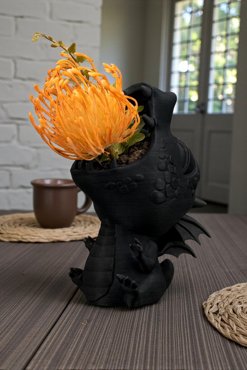 May include: A black dragon-shaped planter with an open mouth, holding an orange flower. The dragon has detailed scales and wings. The flower is a vibrant orange color. The planter is sitting on a wooden table.