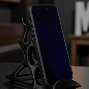 May include: A black smartphone rests on a unique, stylized phone stand. The stand features an intricate, organic design with a curved base and supporting structure. The phone's screen is dark, contrasting with the stand's matte finish. The stand is designed to hold a phone upright.