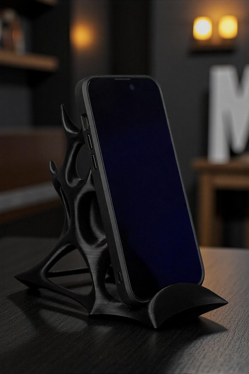 May include: A black smartphone rests on a unique, stylized phone stand. The stand features an intricate, organic design with a curved base and supporting structure. The phone's screen is dark, contrasting with the stand's matte finish. The stand is designed to hold a phone upright.