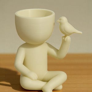 Op de afbeelding: Een cr&egrave;mekleurig beeldje van een zittende persoon met gekruiste benen, die een kleine vogel vasthoudt. De figuur heeft een holle kop, ontworpen om kleine planten of voorwerpen in te bewaren. De vogel zit op de uitgestrekte hand van de figuur, wat een grillig decoratief stuk cre&euml;ert.