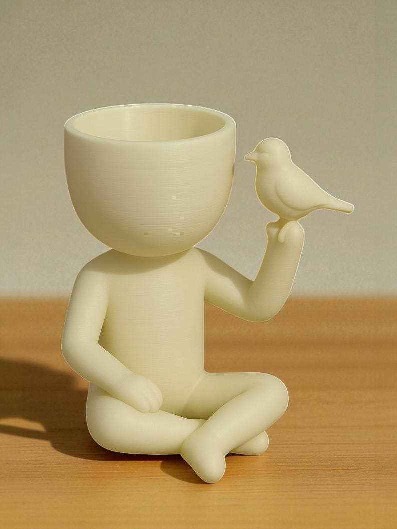 Op de afbeelding: Een cr&egrave;mekleurig beeldje van een zittende persoon met gekruiste benen, die een kleine vogel vasthoudt. De figuur heeft een holle kop, ontworpen om kleine planten of voorwerpen in te bewaren. De vogel zit op de uitgestrekte hand van de figuur, wat een grillig decoratief stuk cre&euml;ert.