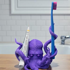Puede incluir: Un soporte para cepillo de dientes con forma de pulpo morado con un cepillo de dientes azul y un tubo de pasta de dientes. El cepillo de dientes tiene un cabezal de cerdas rosa y azul. El soporte est&aacute; sobre una superficie de madera en un ba&ntilde;o.